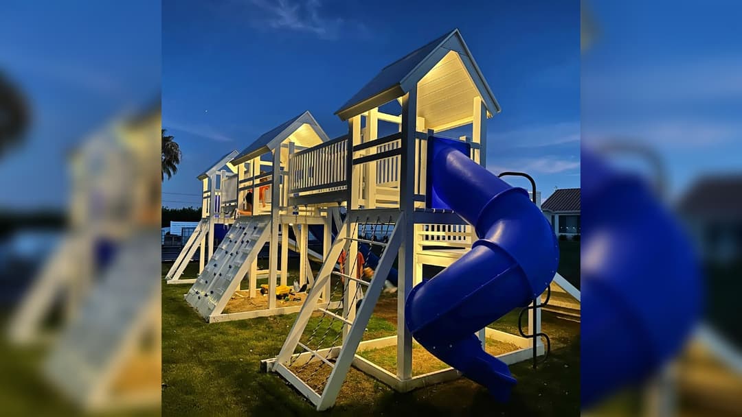 Large white wooden backyard playset with blue spiral slide, climbing walls, and lit towers at dusk.