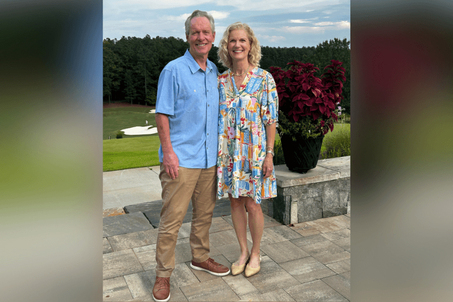Smiling couple posing on a stone patio overlooking a green golf course, with a potted red plant beside them.