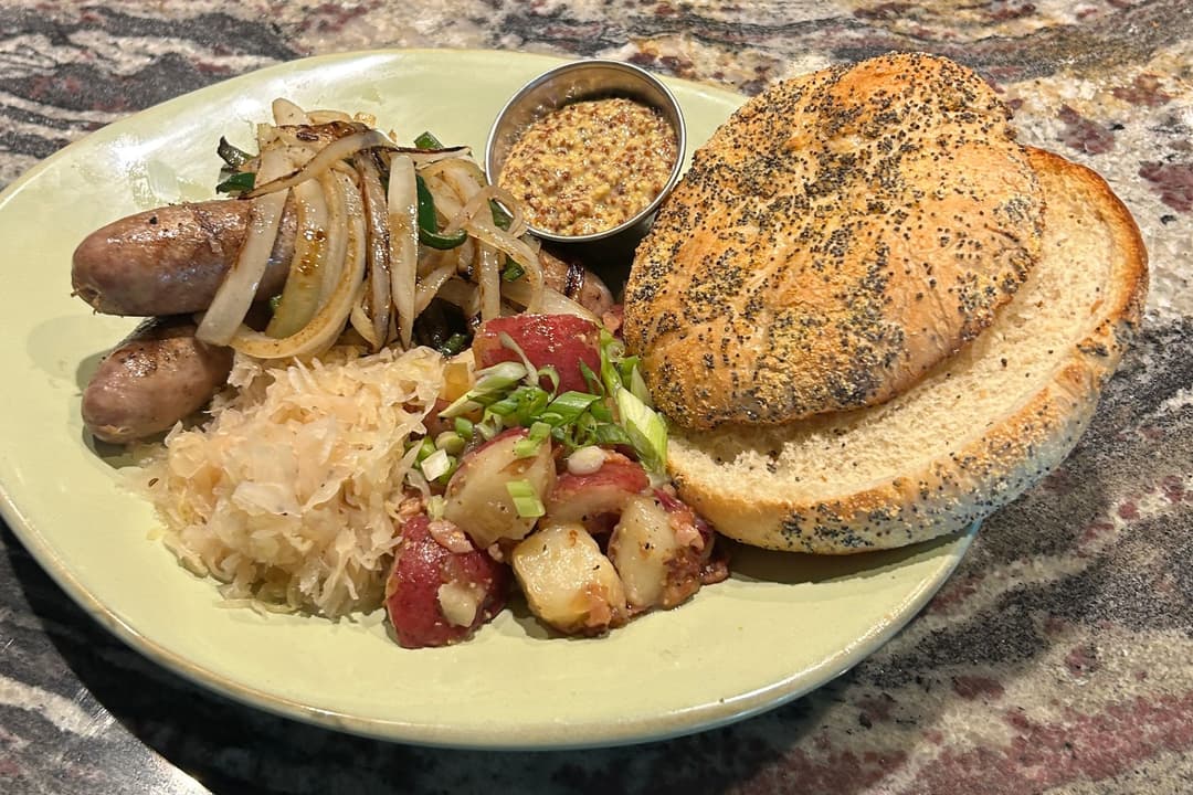 Grilled bratwurst with onions, sauerkraut, potato salad, poppy seed roll, and mustard on a green plate.
