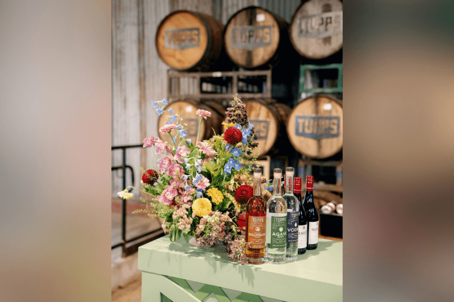 Tupps spirit bottles and a floral bouquet on a green table in front of stacked Tupps barrels.