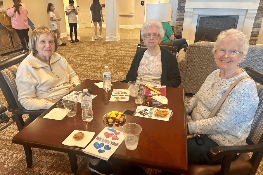Three elderly women sit at a table with snacks and handmade "We Love You" cards in a care facility lounge.