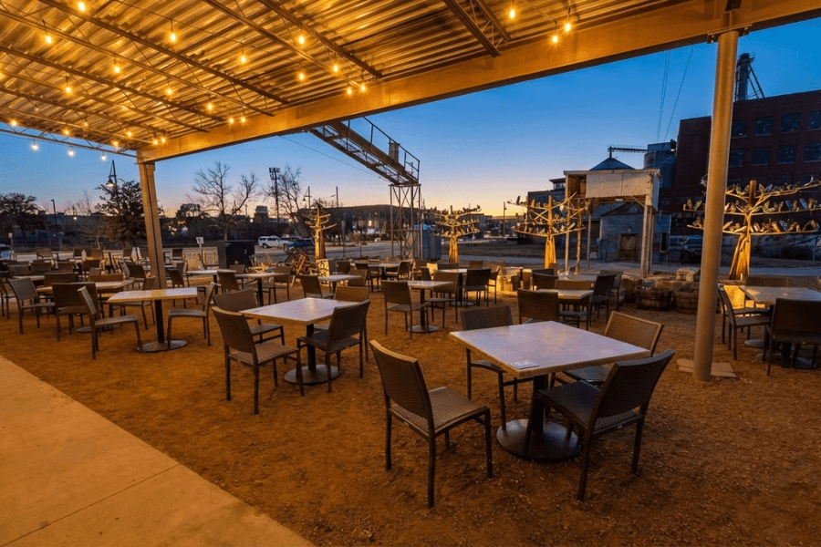 Empty outdoor patio with tables, chairs, and string lights at dusk, with industrial buildings in the background.