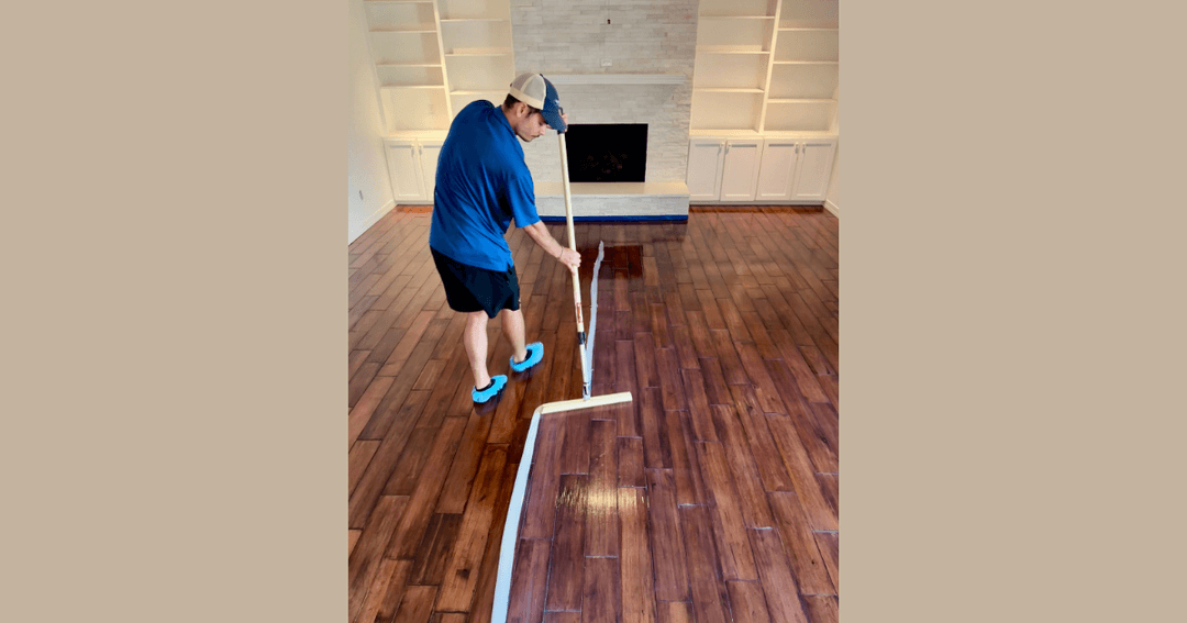 Man applying finish to hardwood floor with an applicator in an empty room with a fireplace and built-in shelves.