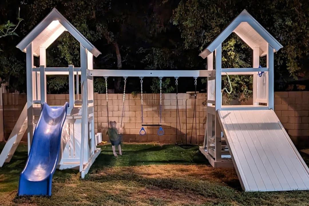 Child on a swing in a backyard playset at night, with blue slide, trapeze bar, and two towers.
