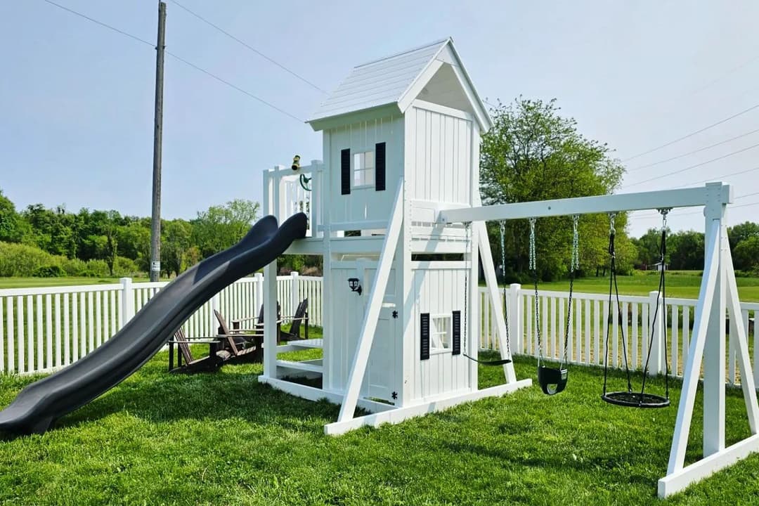 White playhouse-style swing set with black slide, swings, and net seat on a grassy fenced yard.