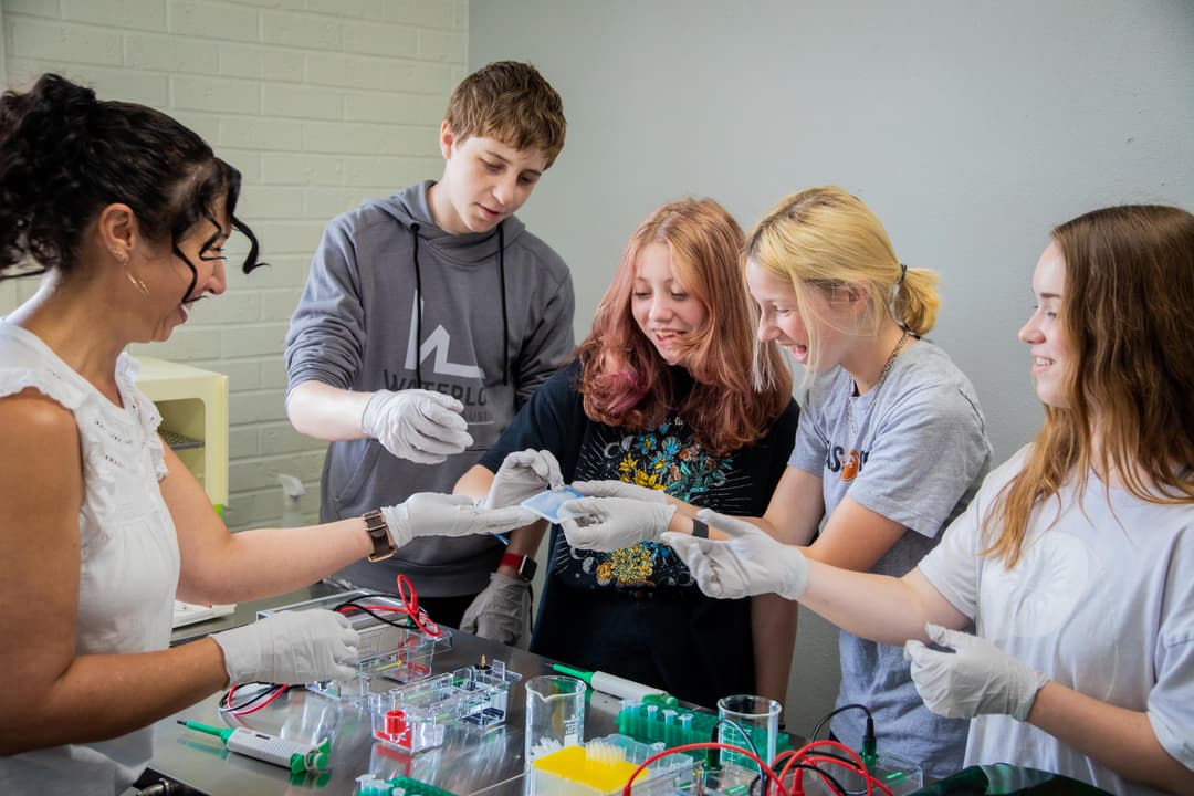 Instructor and four smiling teens in gloves do a gel electrophoresis lab experiment together.