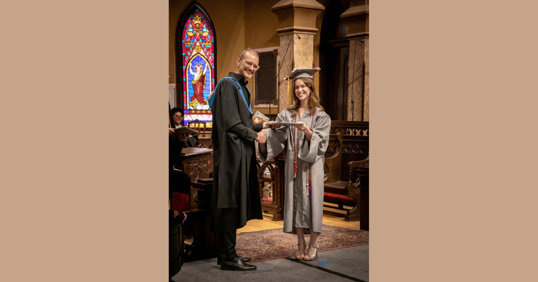 Graduate in cap and gown receives diploma from faculty member in church with stained glass window.