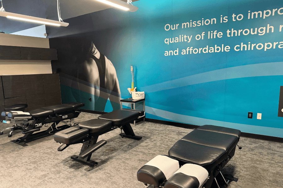 Chiropractic office with adjustment tables, spine model, and teal wall displaying mission statement about affordable care.