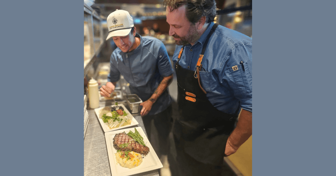 Two chefs plate steak, green beans and mashed potatoes on a stainless steel kitchen counter.