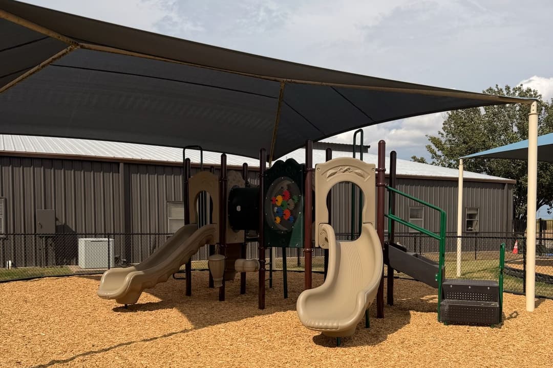 Playground with slides under a dark shade canopy, surrounded by wood chips near a metal building.