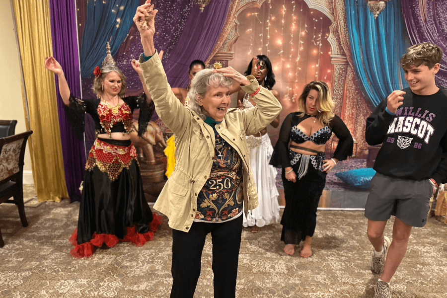 Elderly woman dances with arms raised beside belly dancers and a teen at an indoor celebration.