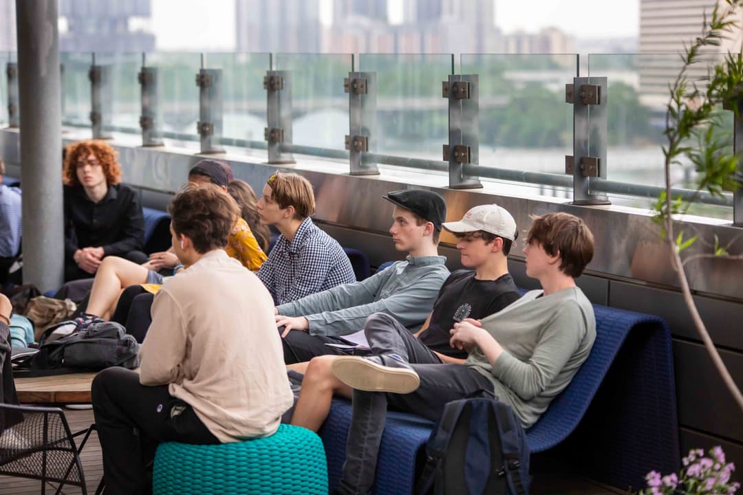 Group of young people seated on a rooftop terrace with a city skyline and river in the background.