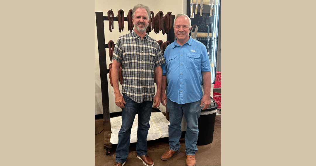 Two men stand smiling in a butcher shop by a rack of hanging smoked sausage links.