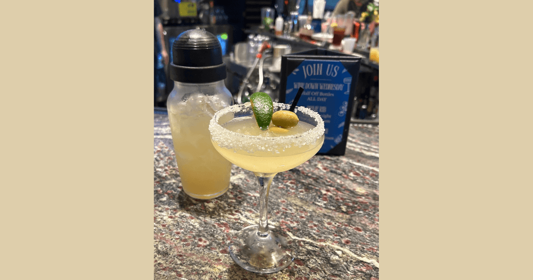 Salt-rimmed margarita with lime and olives beside a cocktail shaker on a bar countertop.