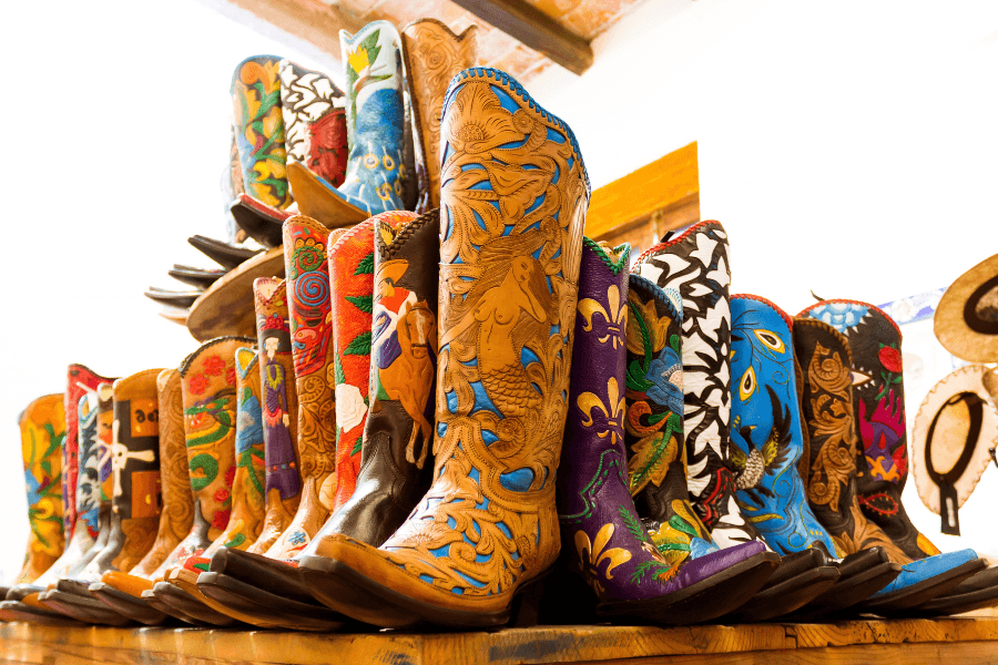 Row of colorful hand-tooled and painted leather cowboy boots displayed on a wooden shelf.