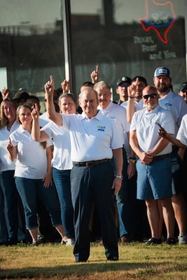 Group in white polo shirts raise index fingers outside a Ford dealership with neon Texas sign.
