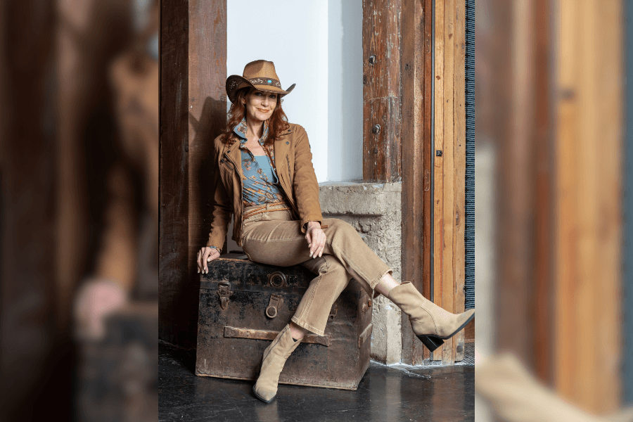 Woman in cowboy hat, tan jacket and boots sits on a vintage trunk against a rustic wood wall.