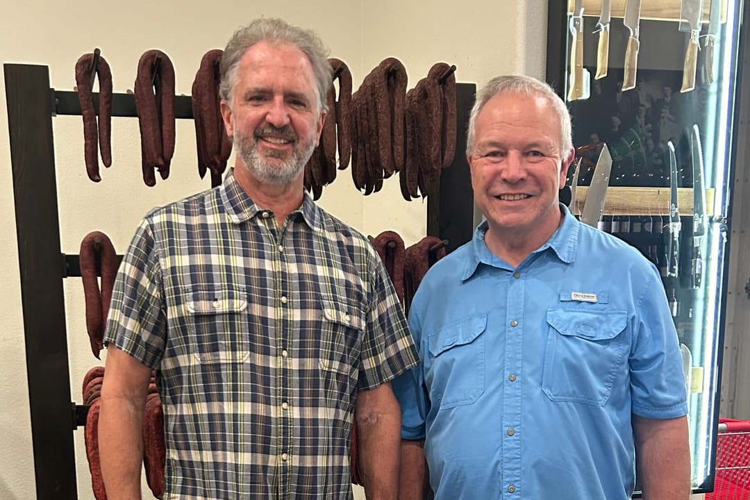 Two men stand smiling in a butcher shop by a rack of hanging smoked sausage links.