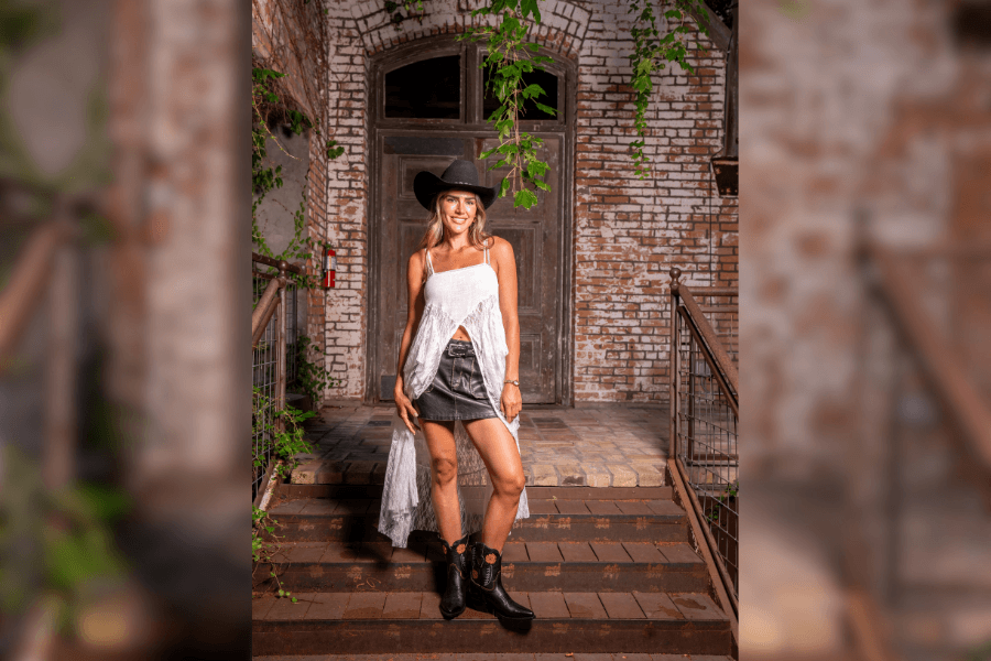 Woman in cowboy hat, white fringe top, denim skirt and boots stands on steps before a rustic brick archway.