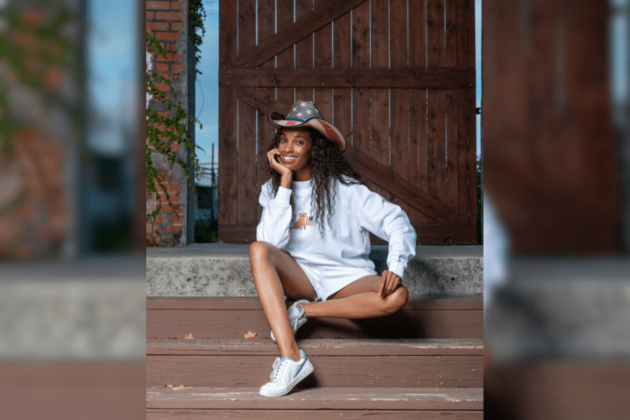 Smiling woman in American flag cowboy hat and white sweatshirt sits on steps by a wooden barn door.