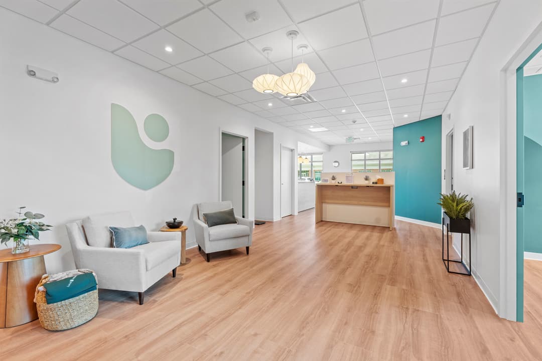 Modern clinic waiting area with white armchairs, wood floors, reception desk and teal accent wall.