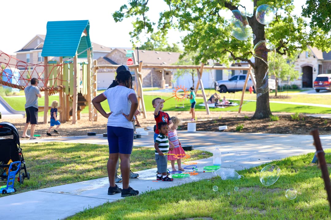 Adults and children play with bubbles at a neighborhood playground with climbing structures and swings on a sunny day.