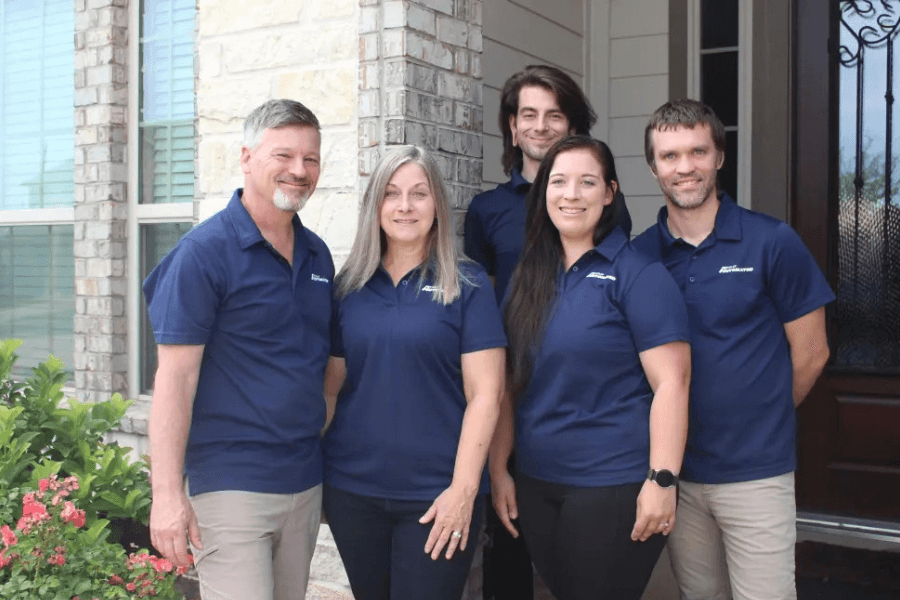 Five people in matching navy polo shirts smile outside a brick house with a wrought-iron door.