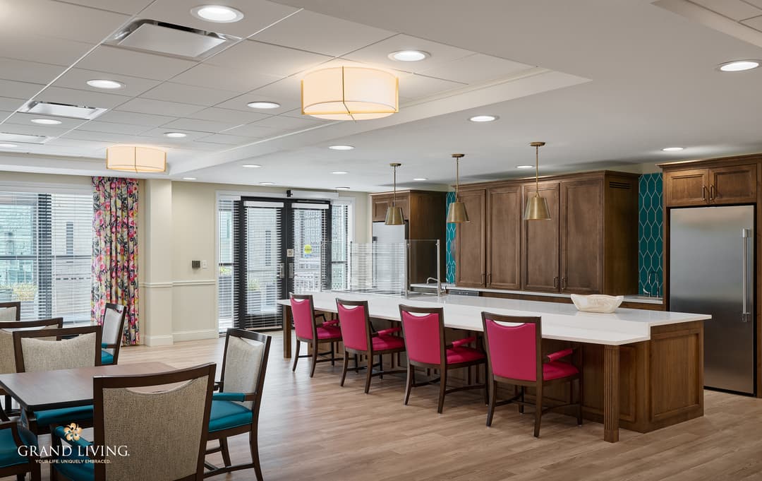 Modern community kitchen with white island, pink bar chairs, wood cabinets, pendant lights, and dining tables.