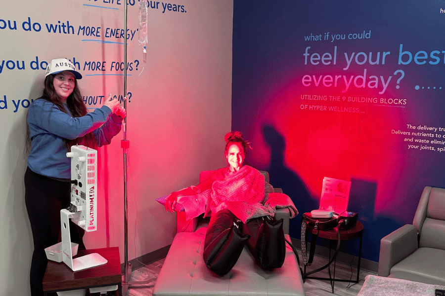 Woman relaxes with IV therapy and compression gear at a wellness spa while staff adjusts equipment under red lighting.