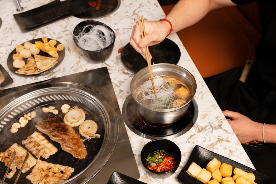 Person uses chopsticks to add noodles to hot pot beside grilled meat, tofu, and dipping sauce at Korean BBQ table.