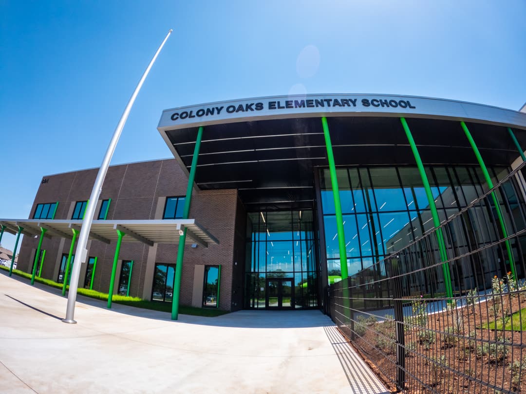 Exterior entrance of modern Colony Oaks Elementary School with green columns, glass facade, and blue sky.