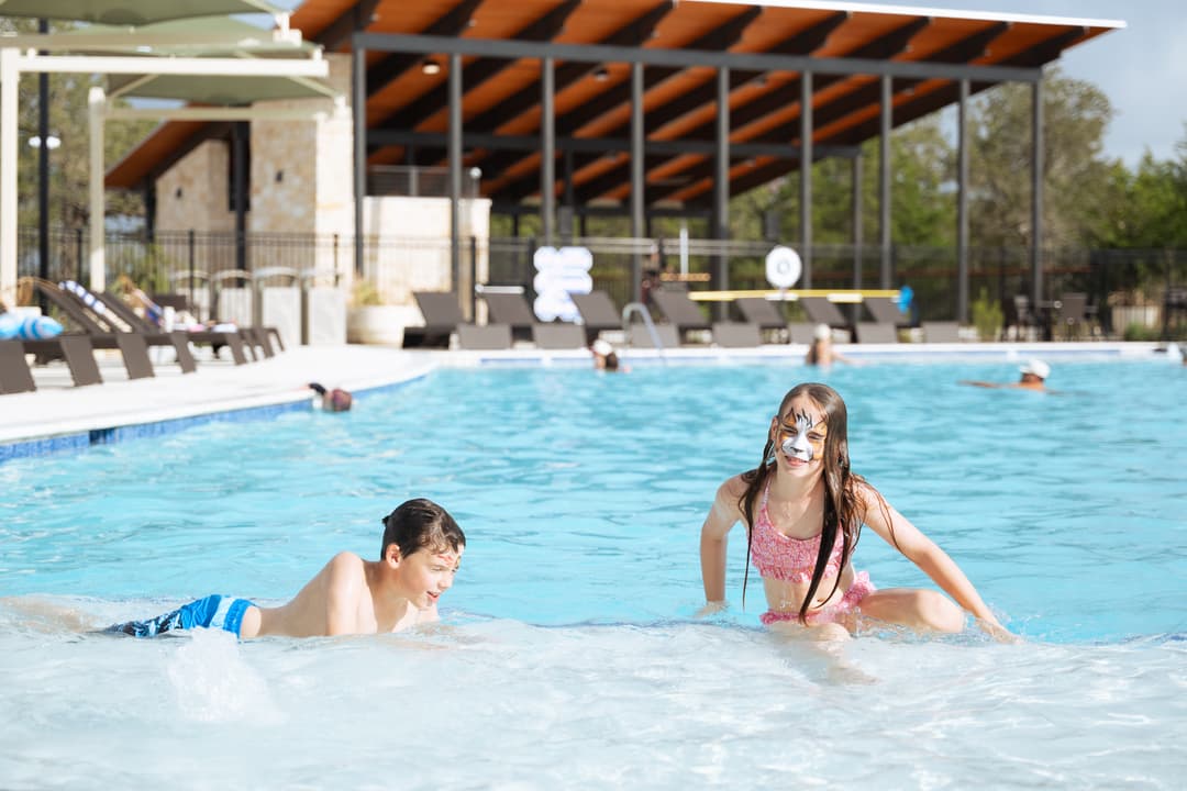 Two children play in shallow pool water, a girl with tiger face paint and a boy, at an outdoor community pool.