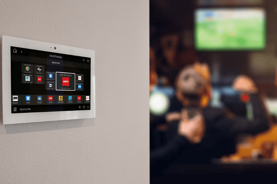 Wall-mounted touchscreen control panel showing sports bar TV channel options, with blurred patrons and a screen behind.