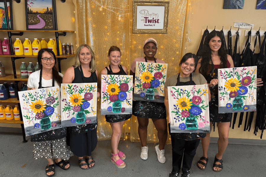 Six women hold up similar floral paintings at a Painting with a Twist studio in Round Rock, Texas.