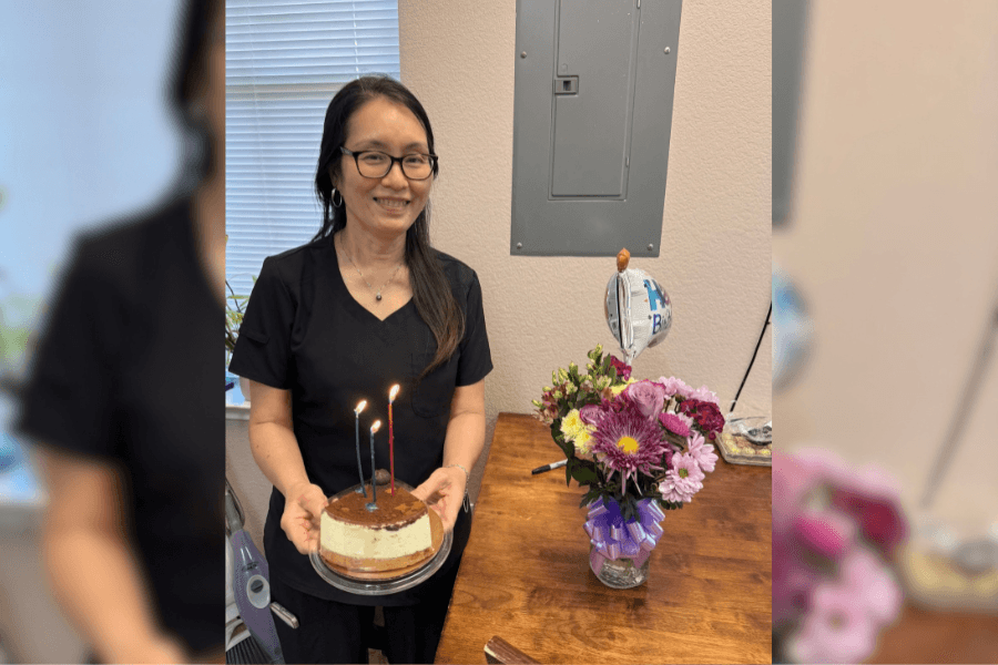 Smiling woman holding a birthday cake with lit candles beside a bouquet of flowers and a balloon.