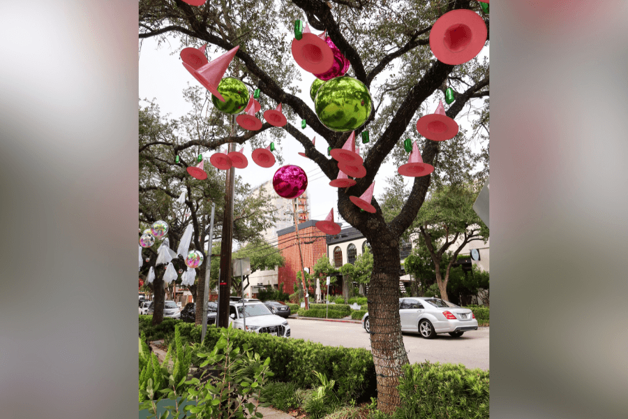 Pink cones and shiny ornaments hang from a tree as public art along a street with parked cars.