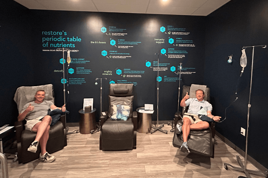 Two men in recliners give thumbs up during IV therapy at a clinic with a "periodic table of nutrients" wall.