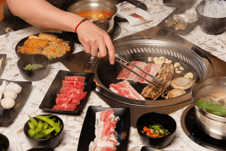 Hand uses tongs to grill sliced meat on a round grill, surrounded by dishes of beef, edamame and dipping sauces.