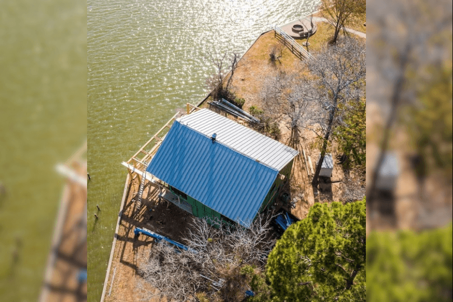 Aerial view of a blue metal-roofed boathouse beside a lake with a dock, trees, and fire pit.