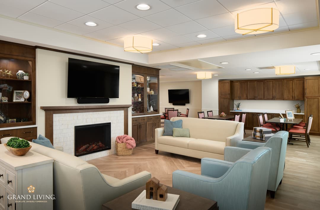 Cozy senior living common room with sofas, armchairs, fireplace, wall-mounted TV, and dining tables beyond.