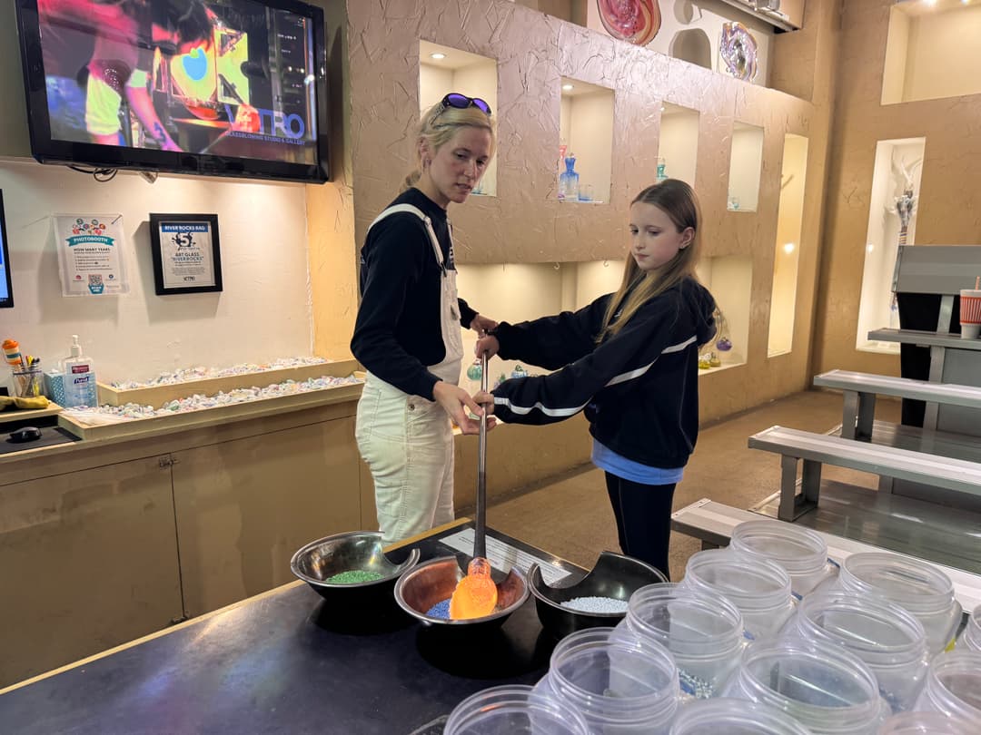 Instructor helps a girl roll molten glass in colored frit at a glassblowing studio.