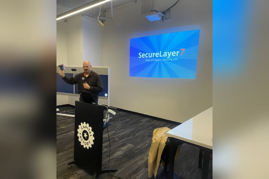 Man speaking at a Capital Factory podium beside a SecureLayer7 presentation slide projected on the wall.