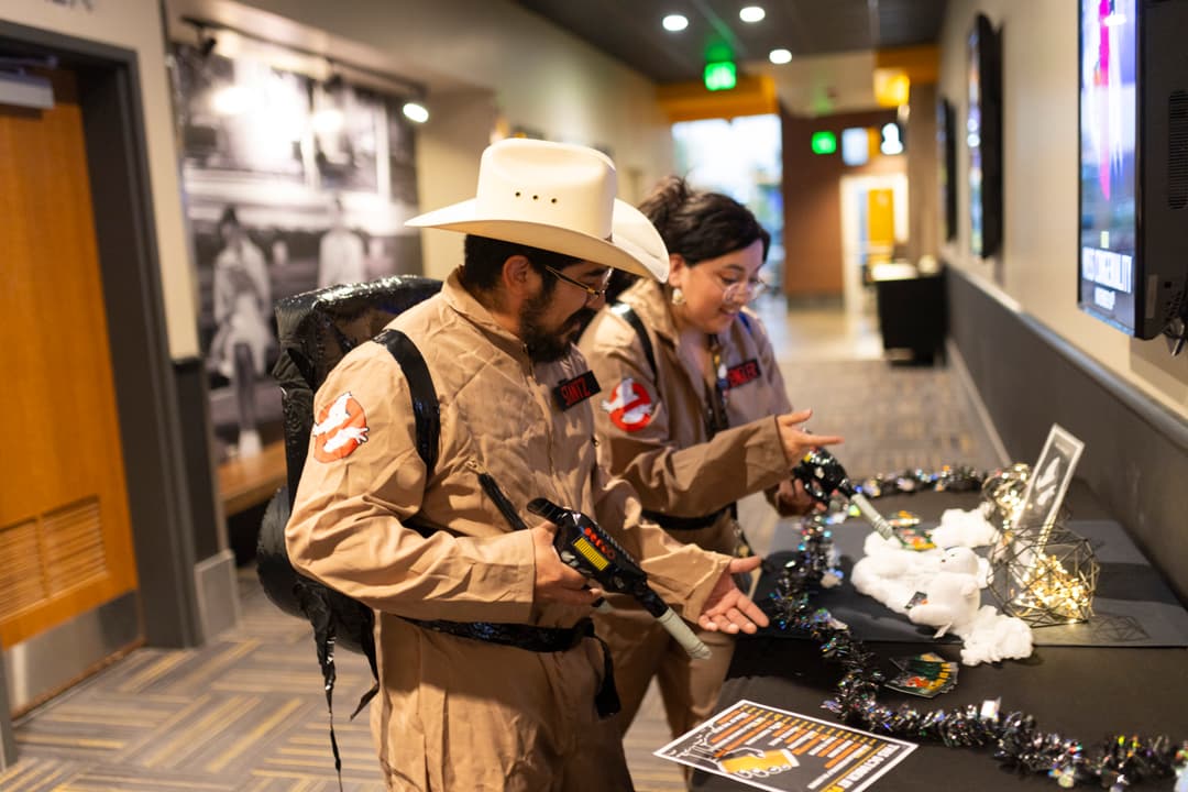 Two people in Ghostbusters costumes with proton packs examine a decorated display table in a theater lobby.
