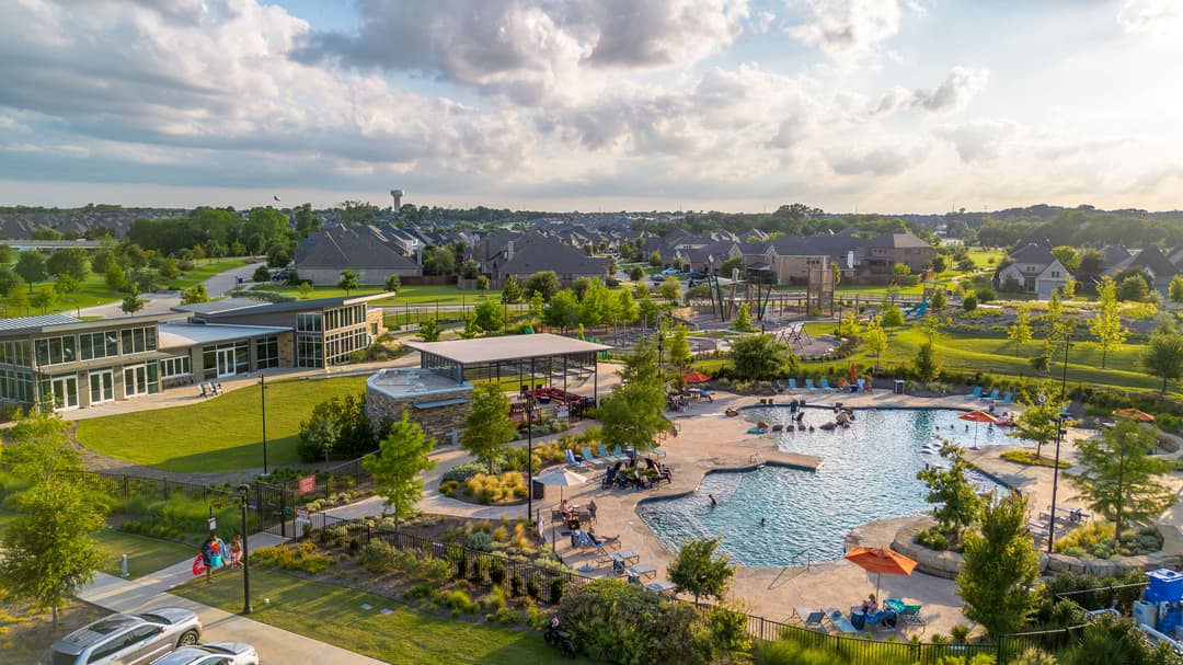 Aerial view of a community pool with orange umbrellas, clubhouse and playground amid a suburban neighborhood with water tower.
