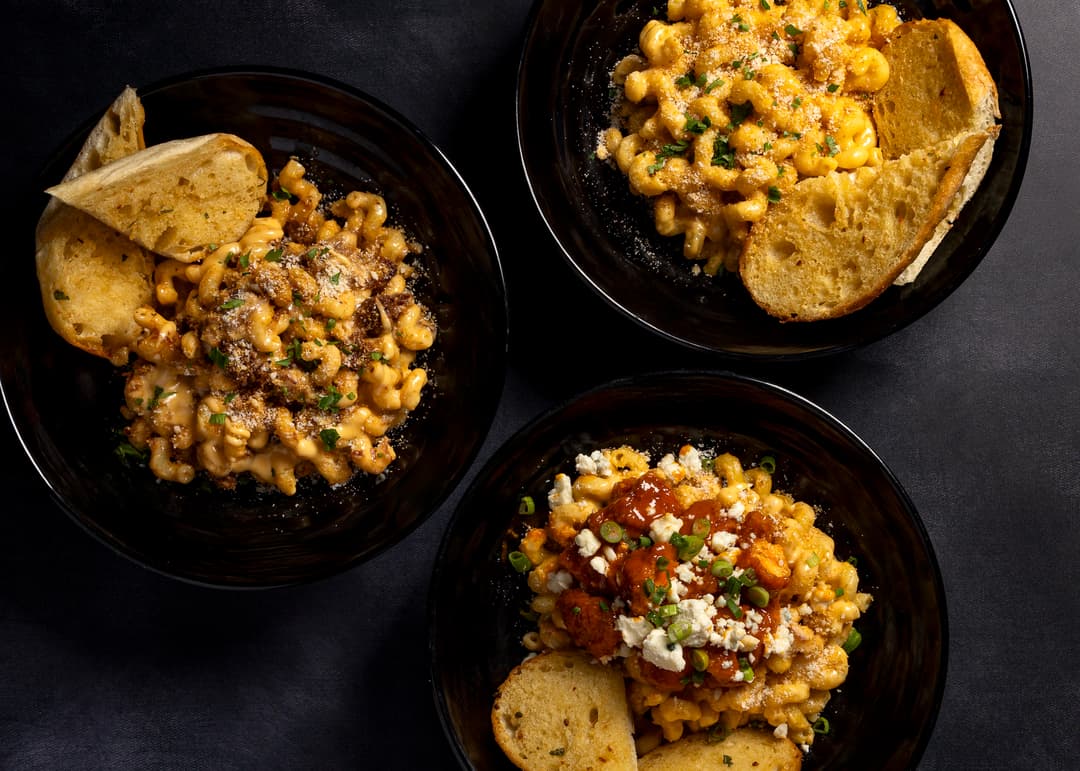 Three black bowls of macaroni and cheese with varied toppings and bread slices on a dark surface.