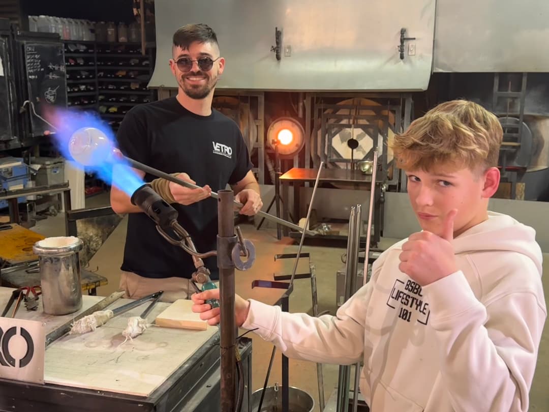 Boy giving thumbs-up while a glassblower heats glass on a rod with a blue flame torch in a studio.