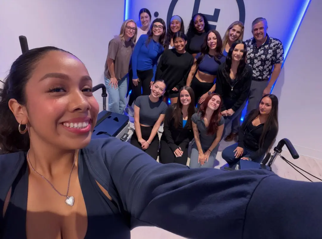 Woman takes a selfie with a smiling group in a fitness studio with Pilates reformers and blue lighting.