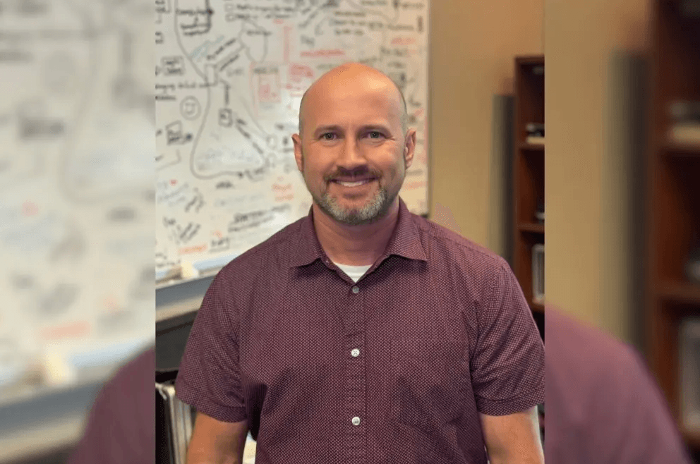 Smiling bald man with goatee in maroon shirt stands in office before a whiteboard with handwritten notes.