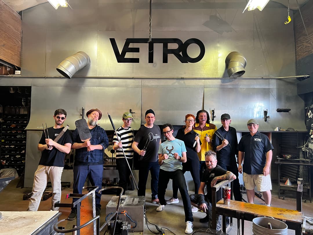 Group of glassblowers holding tools pose under a "VETRO" sign in a glassblowing studio.