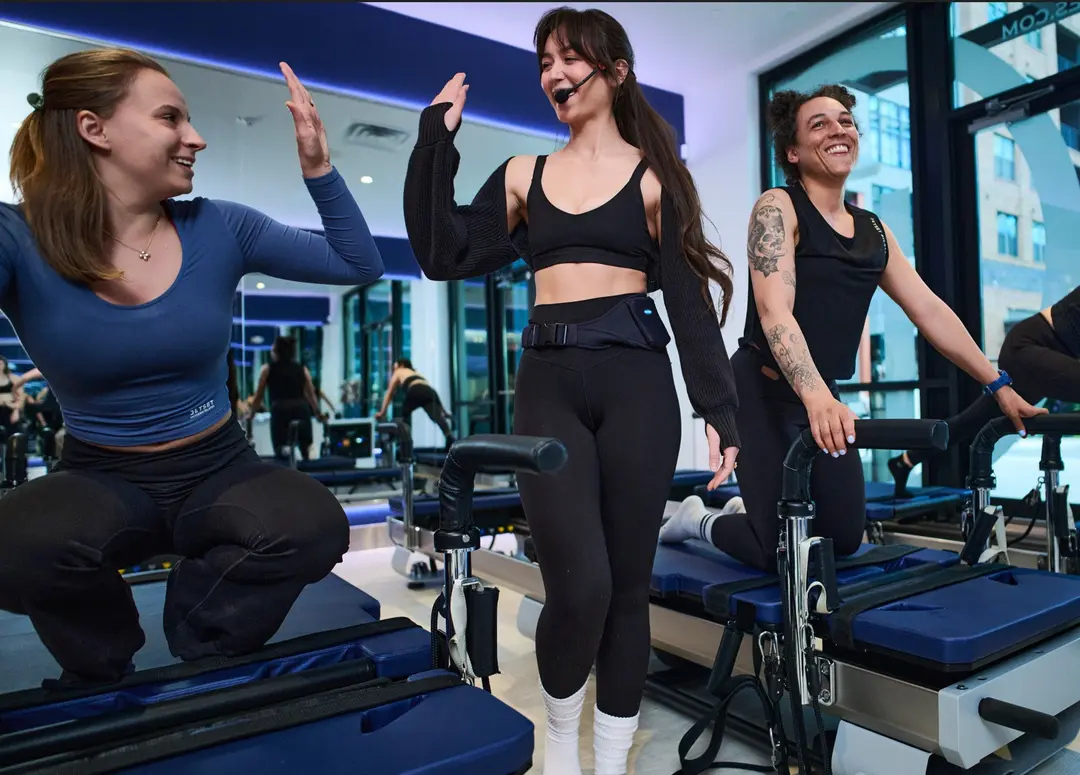 Instructor with headset mic high-fives a smiling woman at a Pilates reformer studio as another participant looks on.